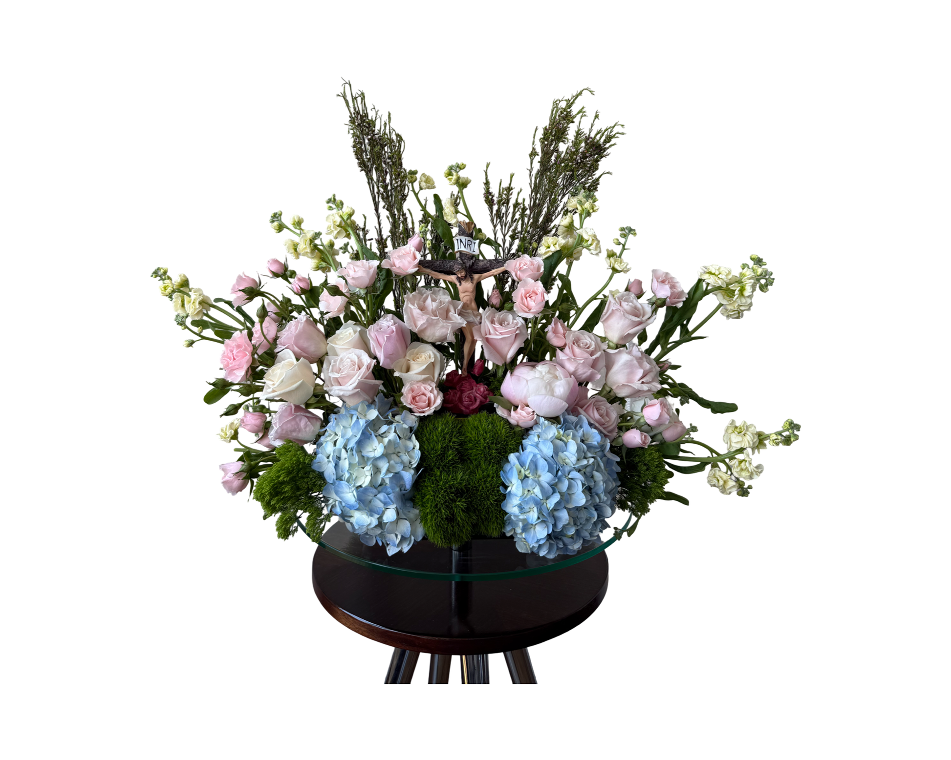A religious-themed floral arrangement featuring a crucifix at its center, surrounded by blush pink and ivory roses, baby pink spray roses, blue hydrangeas, white stock, green moss, and airy fillers. Designed on a dark pedestal base, this display conveys faith, serenity, and reverence—ideal for memorials, funerals, and religious ceremonies.