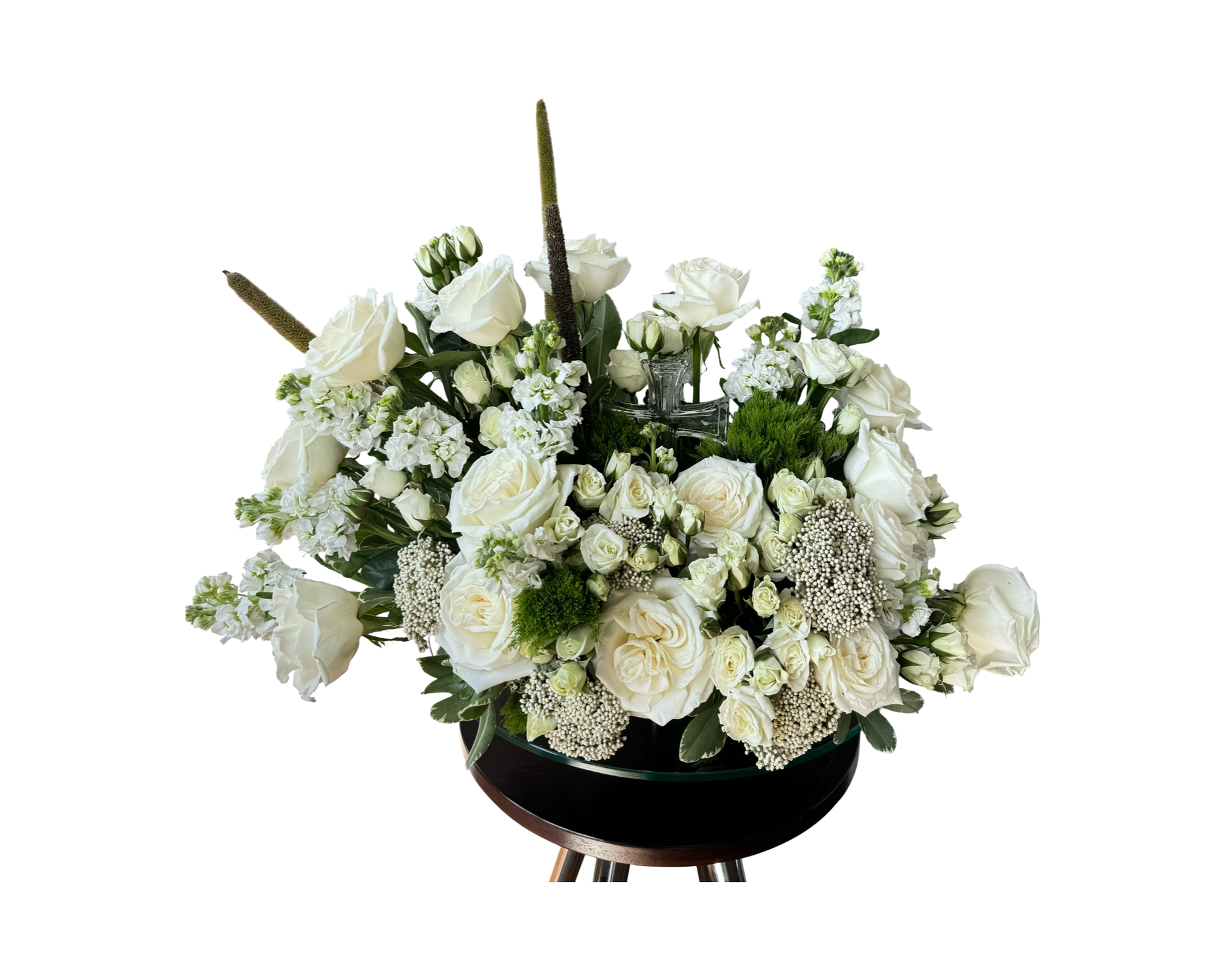A graceful sympathy or ceremonial floral arrangement featuring white roses, white spray roses, white stock, Queen Anne’s lace, green moss accents, and vertical millet spikes. Arranged in a round glass container on a dark pedestal, the piece is balanced and serene—ideal for memorials, religious services, or formal sympathy gifting.
