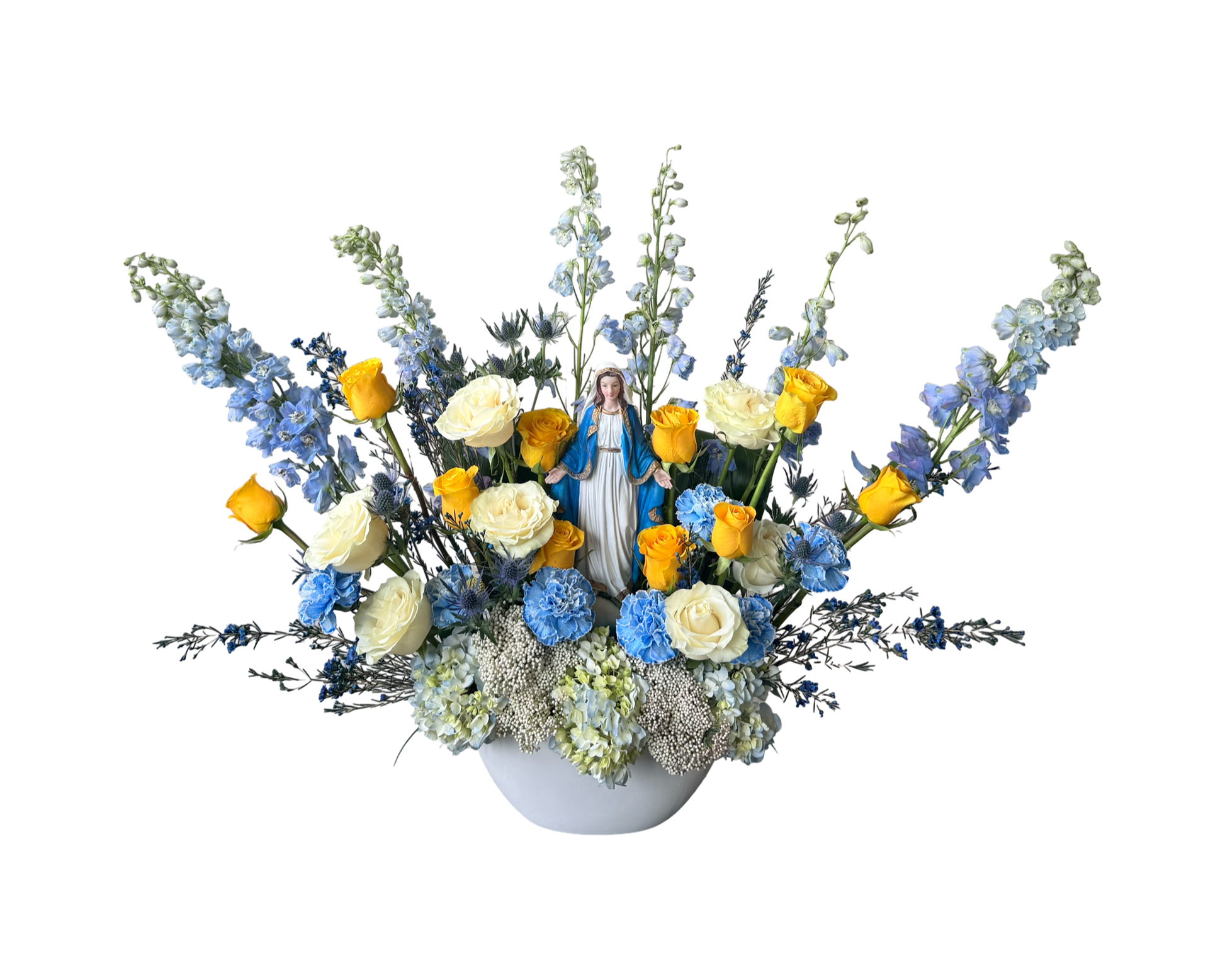 A religious-themed floral arrangement featuring a central Virgin Mary statue surrounded by yellow roses, ivory roses, blue carnations, blue delphinium, blue hydrangeas, yellow craspedia, Queen Anne’s lace, and soft greenery. The celestial blue and golden yellow palette evokes peace, purity, and faith—ideal for church events, funerals, or religious ceremonies.