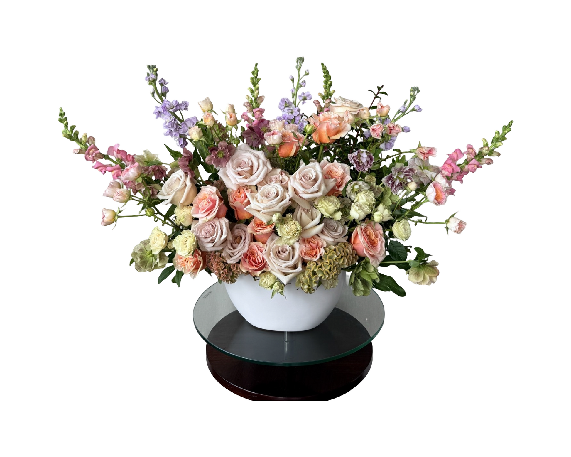 A romantic garden-style floral arrangement in a white ceramic bowl vase, displayed on a glass pedestal. This bouquet features blush and nude roses, peach spray roses, green carnations, lavender stock, soft pink snapdragons, and delicate lisianthus. The airy and pastel color palette makes it ideal for weddings, sympathy tributes, or graceful celebrations.