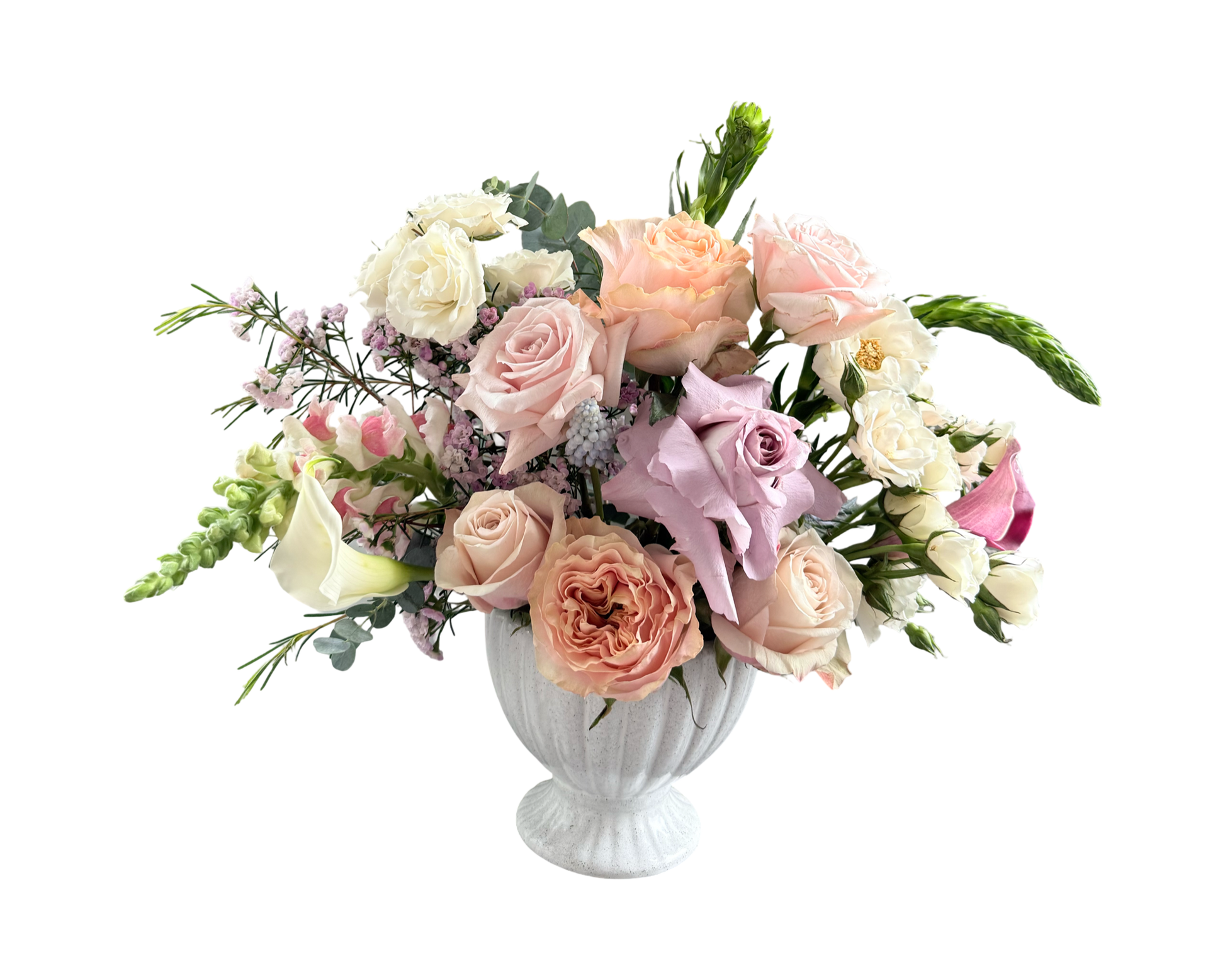 A romantic garden-style floral arrangement in a white fluted compote vase featuring blush roses, peach garden roses, ivory spray roses, lavender roses, white lisianthus, pink calla lilies, snapdragons, pink waxflower, and filler greenery. The pastel palette and classic silhouette make it ideal for weddings, anniversaries, and elegant occasions.