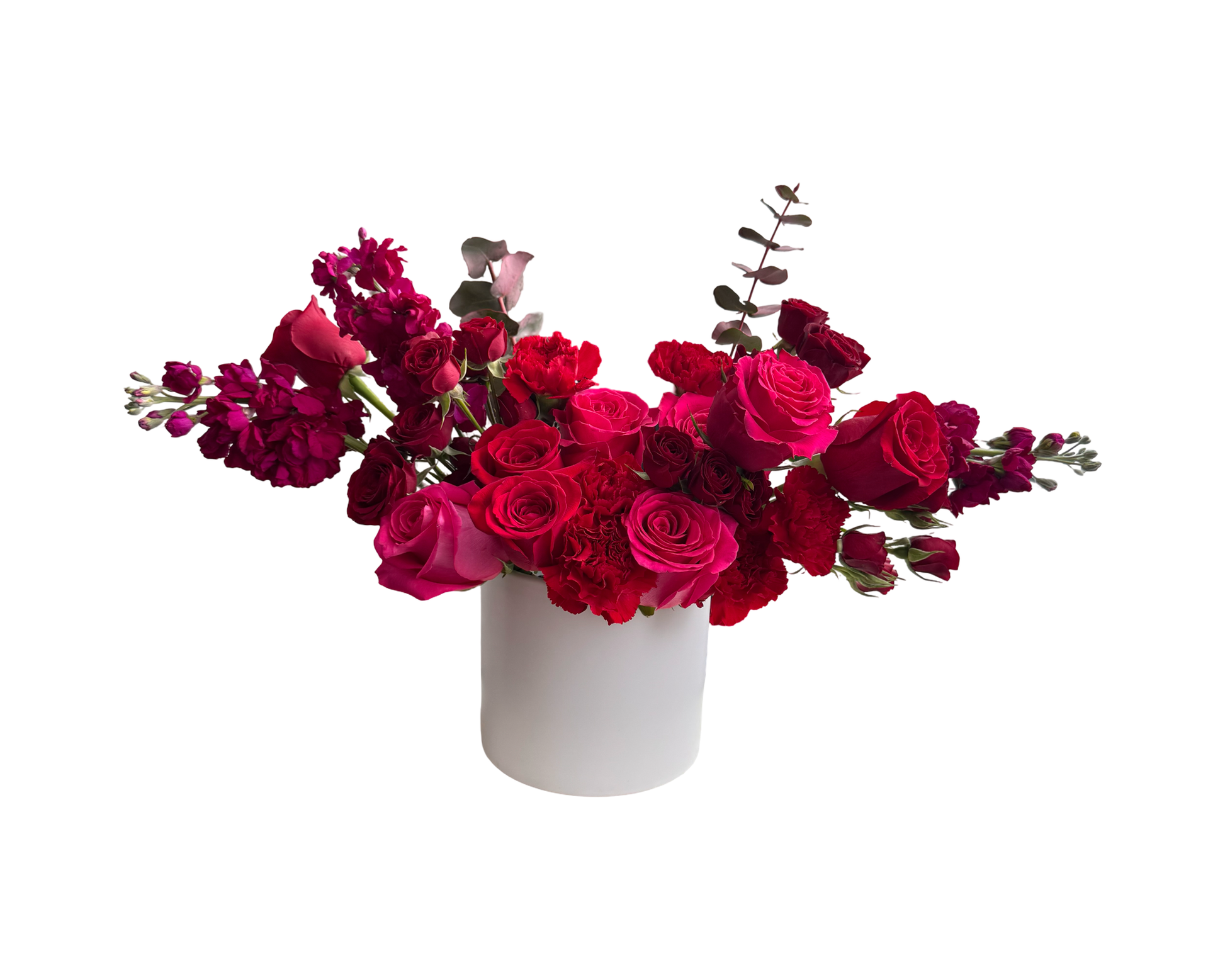 A bold and modern floral arrangement in a white cylindrical vase featuring an elegant mix of fuchsia roses, red roses, red spray roses, red carnations, burgundy stock, and burgundy eucalyptus. The deep jewel tones and varied floral textures create a dramatic, passion-filled statement ideal for love-themed occasions and upscale gifting.
