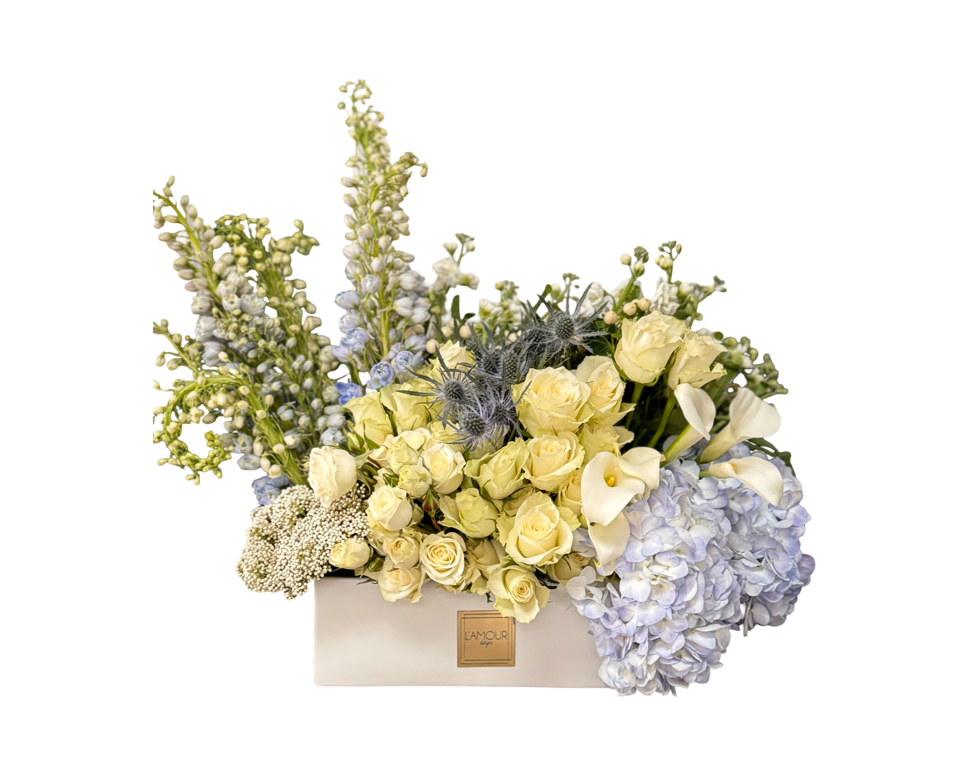 A soft and luxurious floral arrangement presented in a rectangular white ceramic L’amour Designs box, featuring cream roses, spray roses, white calla lilies, blue hydrangeas, blue eryngium (thistle), white delphinium, Queen Anne’s lace, and lush filler greens. This elegant piece offers a peaceful and polished presentation—perfect for sympathy, baptism, or serene celebrations.