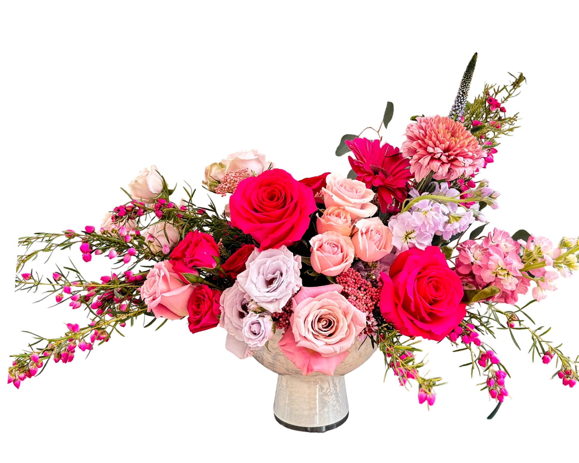 A vibrant floral centerpiece in a rounded marble-like pedestal vase, featuring a striking palette of hot pink, blush, and lavender tones. The arrangement includes bold red roses, soft pink roses, lavender roses, pink waxflower, peach spray roses, mauve chrysanthemums, pink stock, gerbera daisies, and textural filler greenery. A modern-meets-romantic design ideal for celebrations of love, power, and elegance.