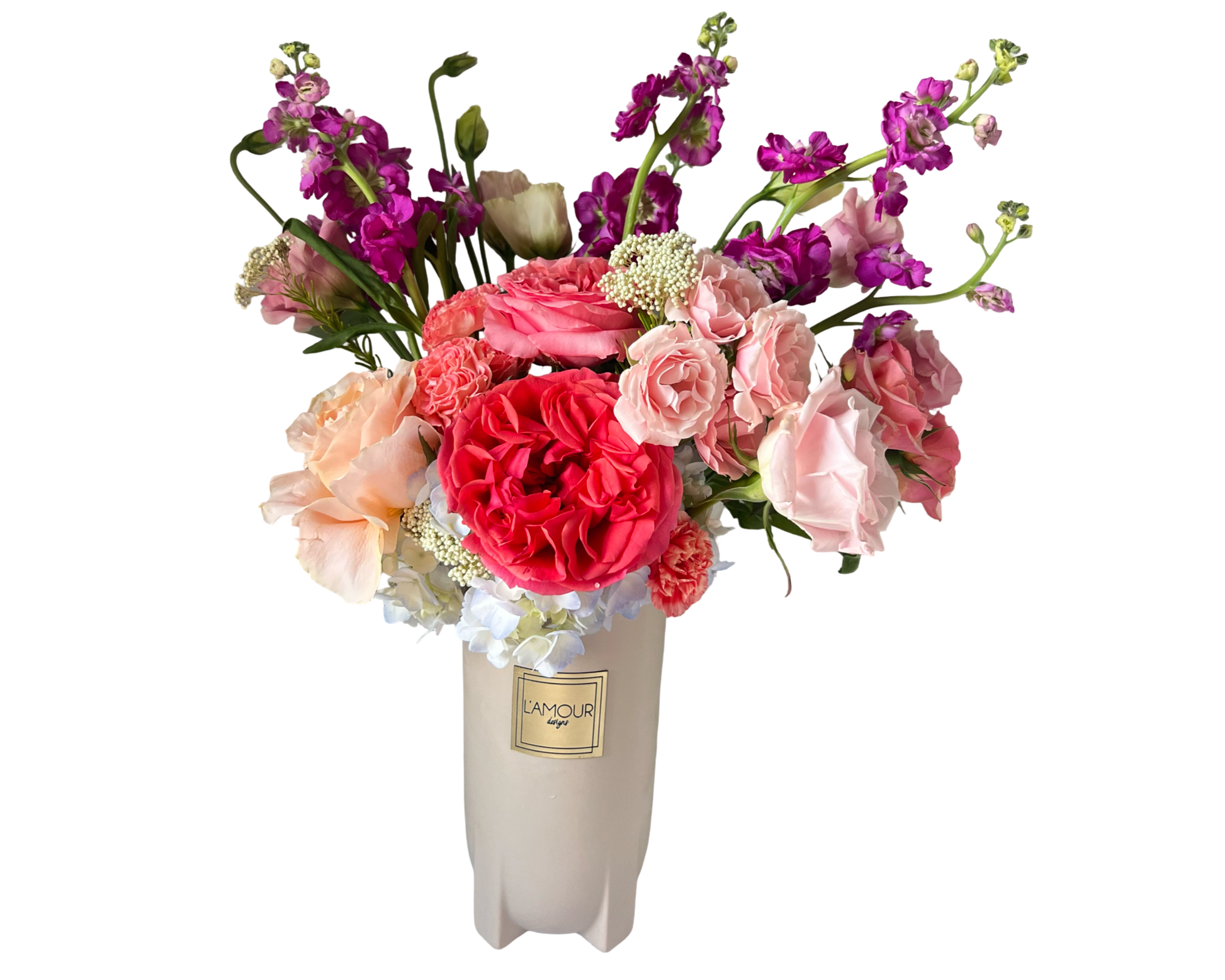 A vibrant and elegant floral arrangement featuring a show-stopping coral garden rose at the center, surrounded by soft pink roses, spray roses, peach roses, and delicate carnations. Accents of white yarrow, airy hydrangeas, and tall purple stock flowers add texture and dimension. Presented in a tall white vase with the L'amour Floral Designs label, this arrangement is perfect for birthdays, anniversaries, Mother’s Day, or as a stunning centerpiece for any celebration.