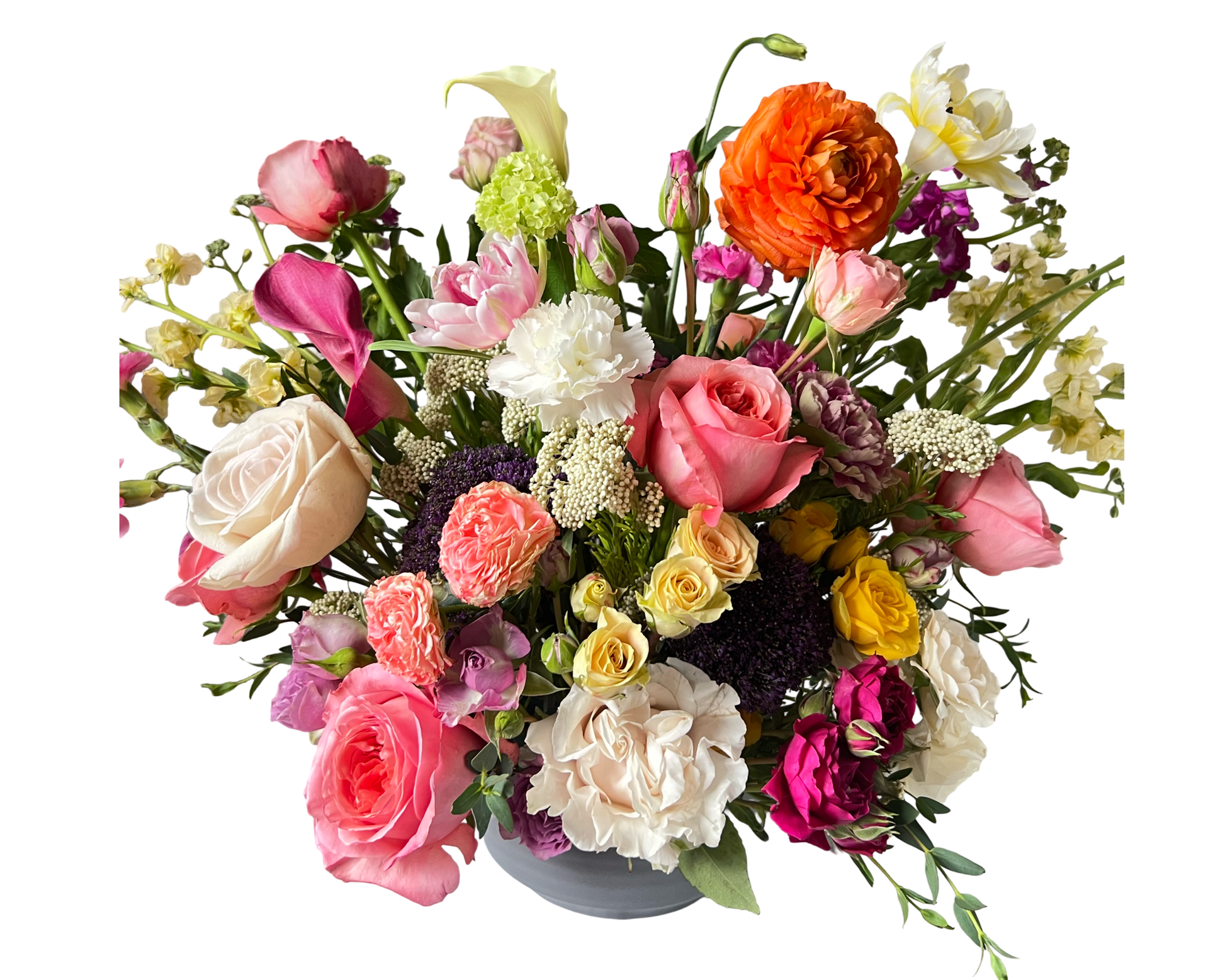 A vibrant and eclectic floral arrangement bursting with a rich variety of blooms including blush pink roses, creamy white roses, cheerful yellow roses, and delicate spray roses. Accents of bright orange ranunculus, elegant calla lilies, carnations, and lisianthus bring texture and depth, while clusters of yarrow and deep purple blooms add contrast. Arranged in a modern white vase, this colorful bouquet is perfect for birthdays, anniversaries, Mother’s Day, or any joyful celebration.