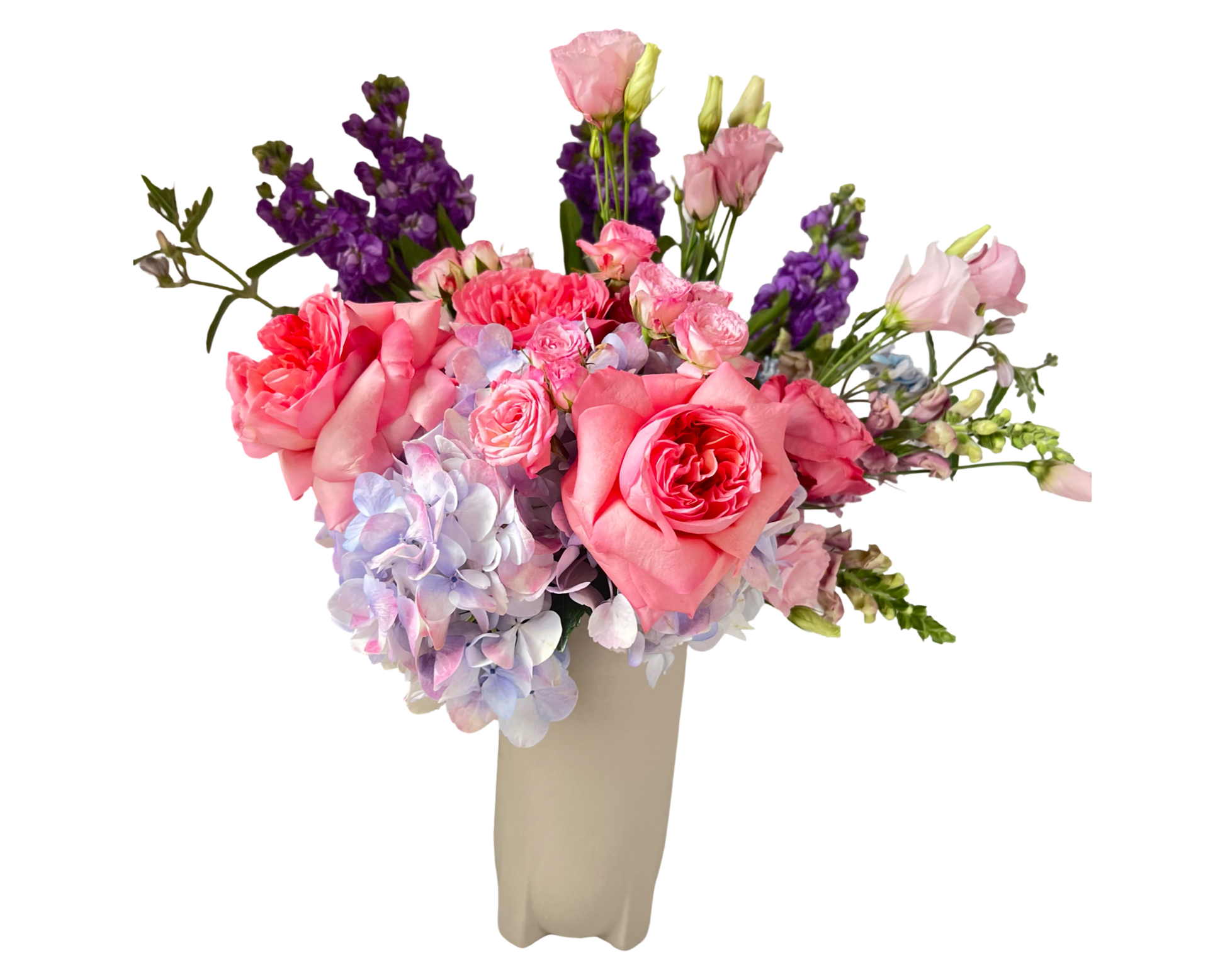 This elegant and feminine floral arrangement is designed in a tall, sculpted beige vase and features a romantic mix of soft pinks, lavenders, and purples. The bouquet includes large garden-style pink roses, pale pink lisianthus, lavender snapdragons, light purple hydrangeas, pink spray roses, and bold purple stock. The blend of textures and gentle tones gives the arrangement a dreamy, graceful feel, perfect for thoughtful occasions, romantic gestures, or celebratory events.