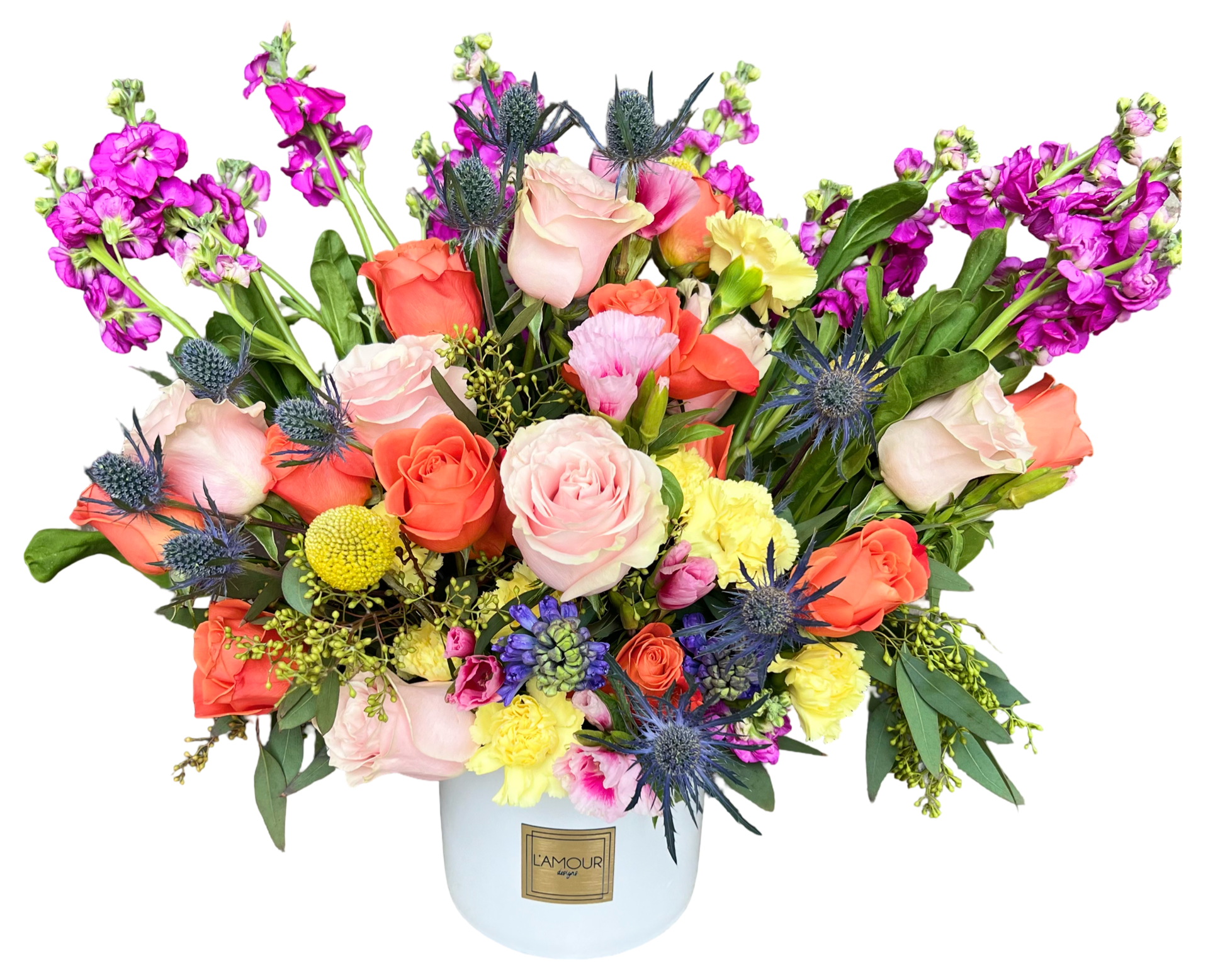 A bold and cheerful floral arrangement in a white ceramic vase by L’amour Designs, filled with a dynamic blend of coral roses, blush roses, yellow carnations, lavender stock, purple eryngium thistle, yellow craspedia, pink spray roses, and lush greenery. This high-contrast bouquet pops with joyful energy and natural texture, perfect for modern celebrations and vibrant gifting.