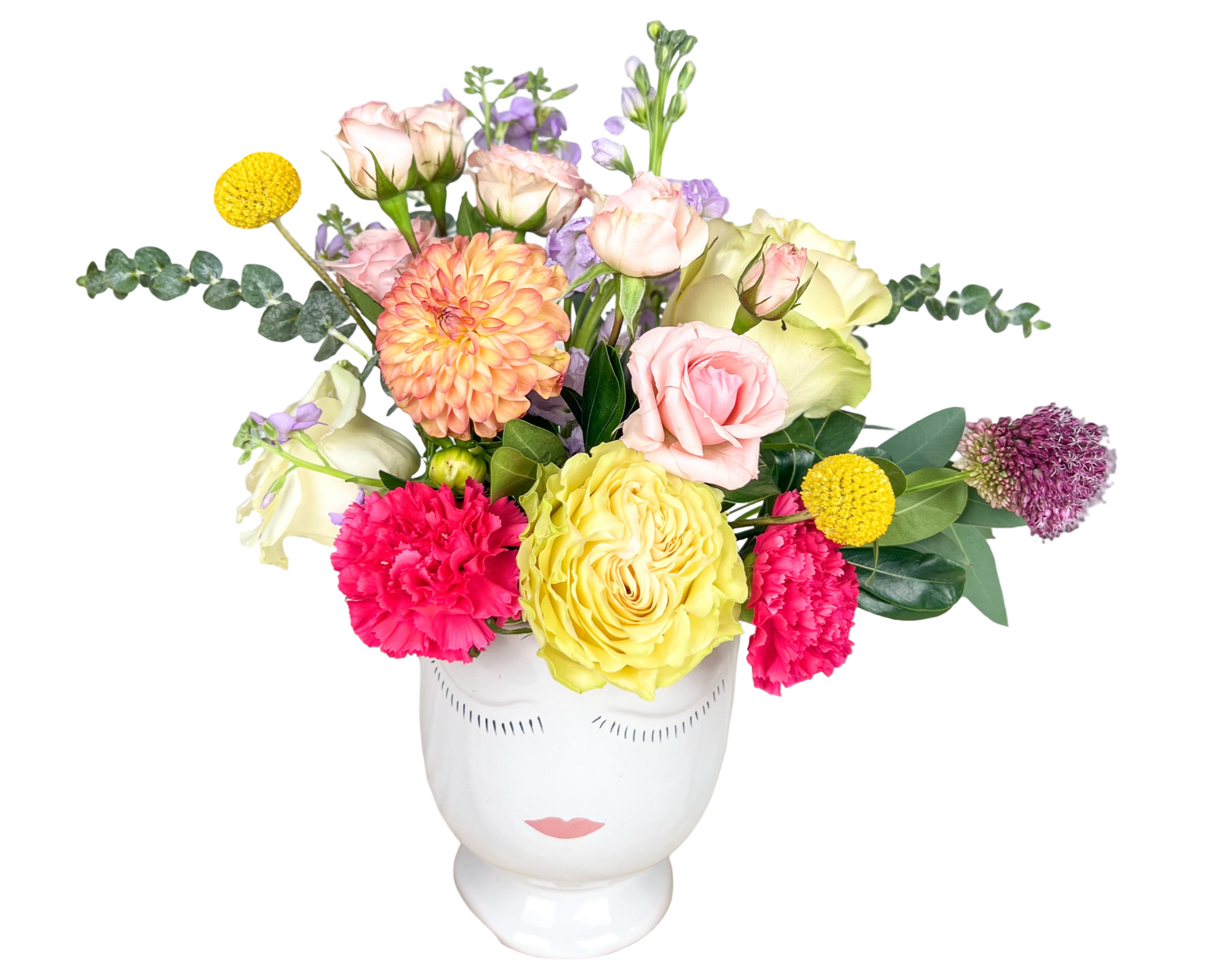 A whimsical and colorful floral arrangement displayed in a white ceramic vase with a serene face design. This bouquet includes soft pink roses, pale yellow garden roses, hot pink carnations, peach dahlias, yellow billy balls (craspedia), lavender stock, eucalyptus stems, and purple allium, accented with seasonal greenery. The design features a playful, artistic aesthetic with a balanced mix of bold and pastel tones—perfect for modern gifting and special occasions.