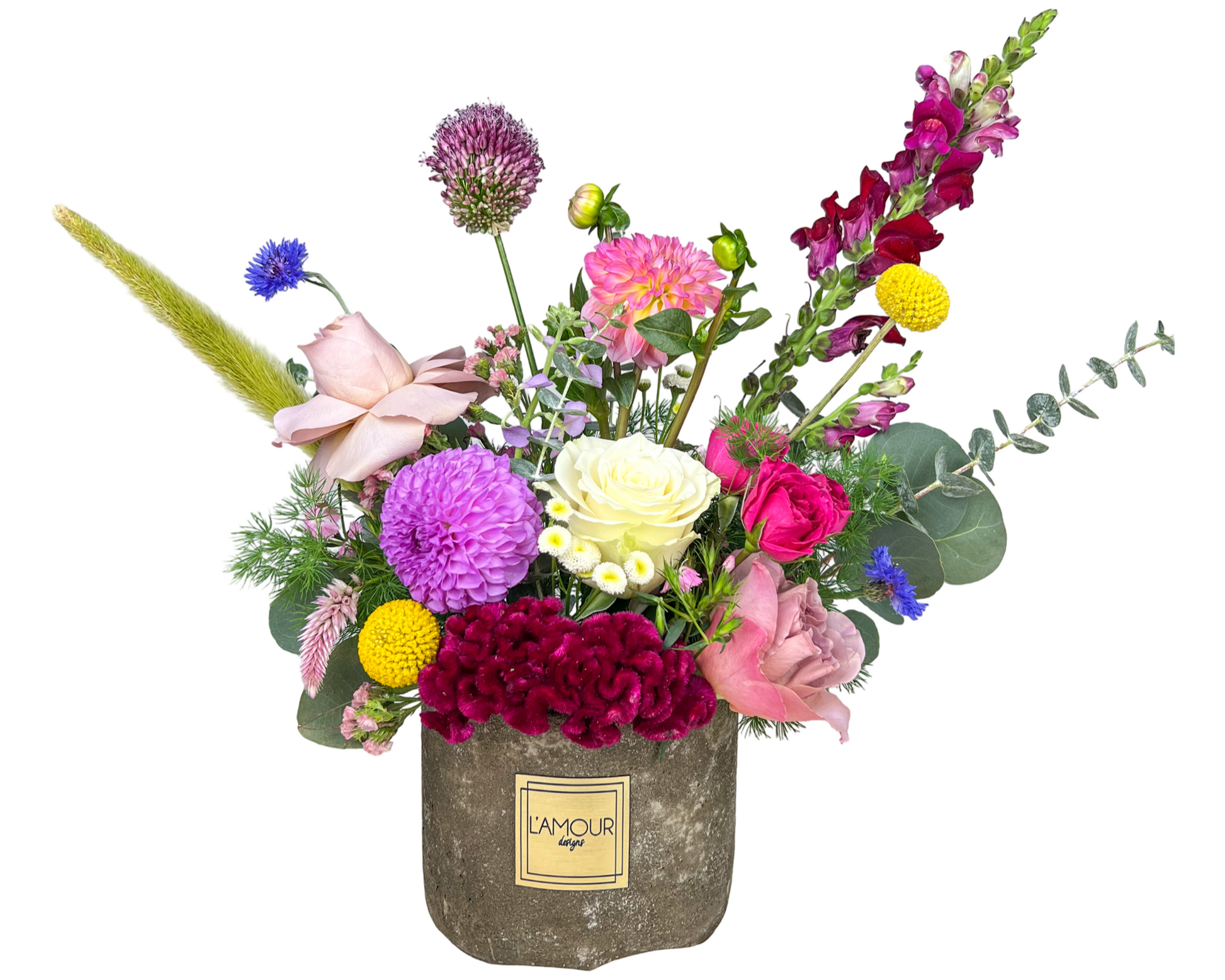 A vibrant and eclectic floral arrangement presented in a rustic stone-textured L’amour Designs container. This design features a striking mix of textures and colors, including white roses, fuchsia spray roses, lavender roses, yellow craspedia (billy balls), purple dahlias, burgundy coxcomb, blue cornflowers, pink lisianthus, burgundy snapdragons, pink dahlias, allium, and various greenery including eucalyptus and grasses. A playful, garden-style piece perfect for creative gifting or joyful occasions.