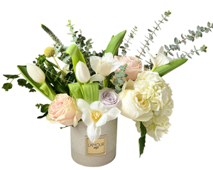 A refined and elegant floral arrangement featuring creamy white hydrangeas, ivory roses, soft blush roses, white tulips, and delicate calla lilies. Hints of pale lavender roses add a subtle touch of color, while eucalyptus and lush greenery provide natural texture and height. Arranged in a modern beige L’Amour vase, this bouquet exudes sophistication and grace, making it ideal for weddings, anniversaries, sympathy tributes, or elegant home décor.