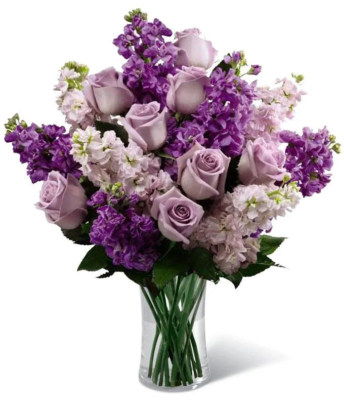 This elegant floral arrangement features a luxurious bouquet of lavender roses surrounded by layers of deep and light purple stock flowers. Set in a clear glass vase, the blooms are densely packed, creating a plush and romantic display. The combination of soft pastel and bold purple hues makes this a perfect statement piece for any elegant setting.