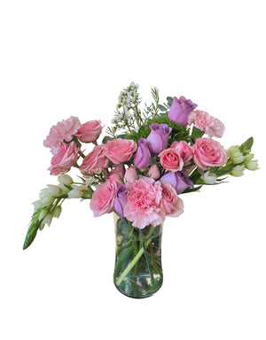 This floral arrangement from L’amour Floral Designs is a cheerful mix of soft pink and lavender roses, light pink carnations, delicate white blooms, and lush greenery displayed in a clear glass vase. The colors are balanced beautifully, evoking a romantic and uplifting tone perfect for springtime occasions or heartfelt sentiments. The arrangement is fresh, airy, and feminine, ideal for expressing love, appreciation, or warm wishes.