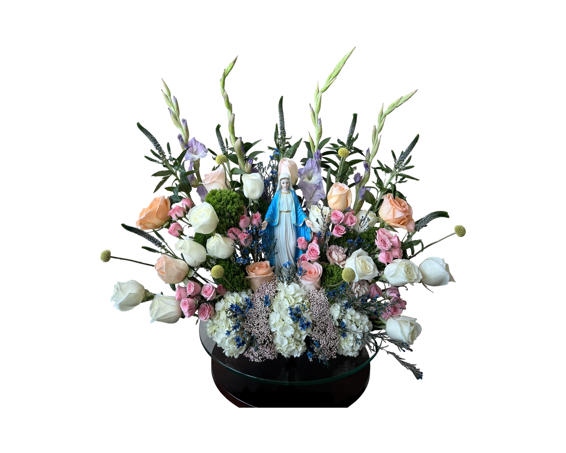 A faith-inspired floral arrangement designed around a ceramic Virgin Mary statue, featuring a pastel mix of ivory roses, peach roses, pink spray roses, white hydrangeas, blue statice, lavender gladiolus, craspedia, moss, and veronica. Displayed on a round glass pedestal, this serene and symbolic display is perfect for religious ceremonies, sympathy tributes, or church decor.
