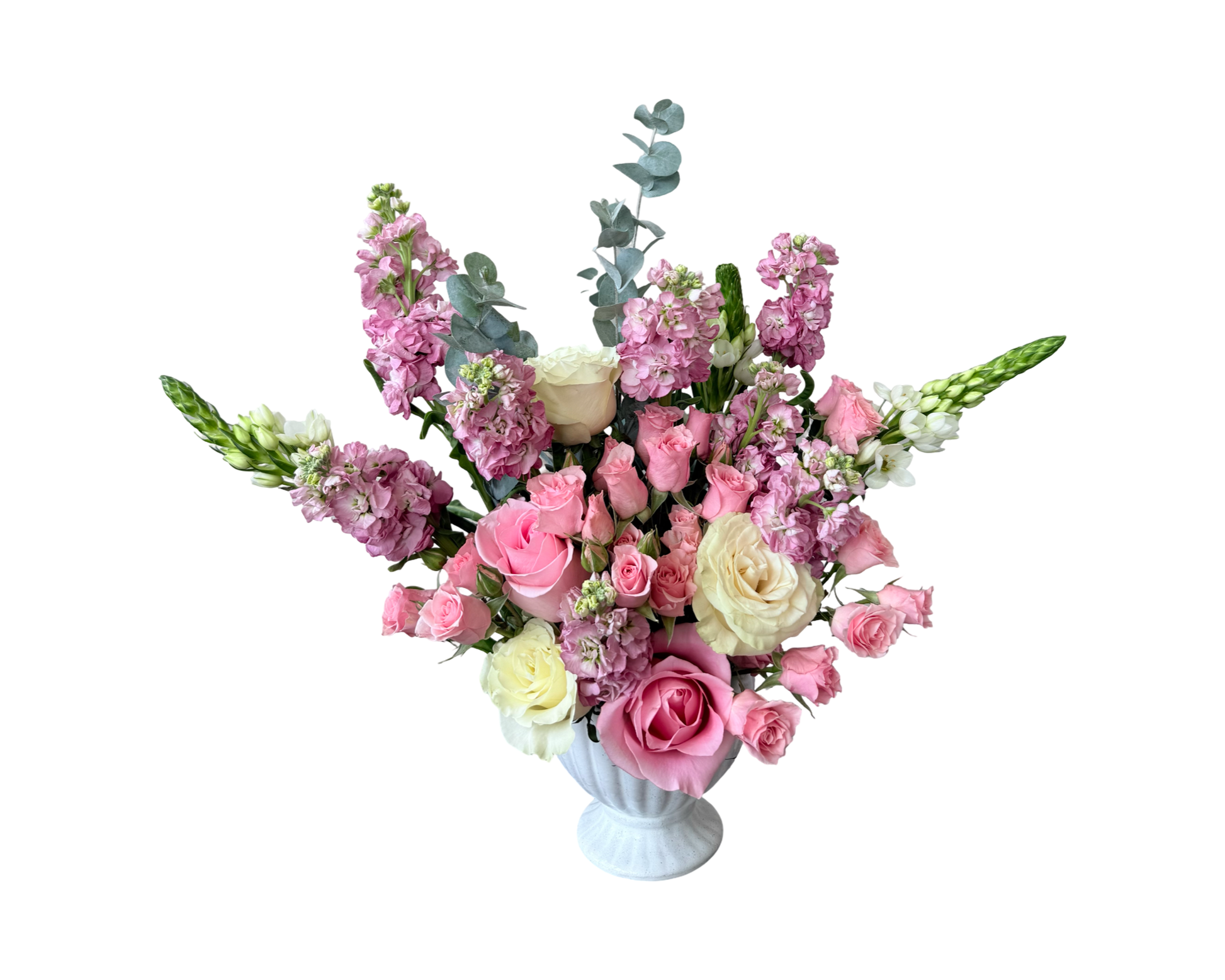 A soft and elegant floral arrangement displayed in a white ceramic fluted urn-style vase. The bouquet features a romantic blend of pale pink roses, ivory roses, blush spray roses, lavender stock, white snapdragons, and eucalyptus stems. The airy, garden-inspired design and pastel tones make it ideal for both classic celebrations and gentle sentiments.