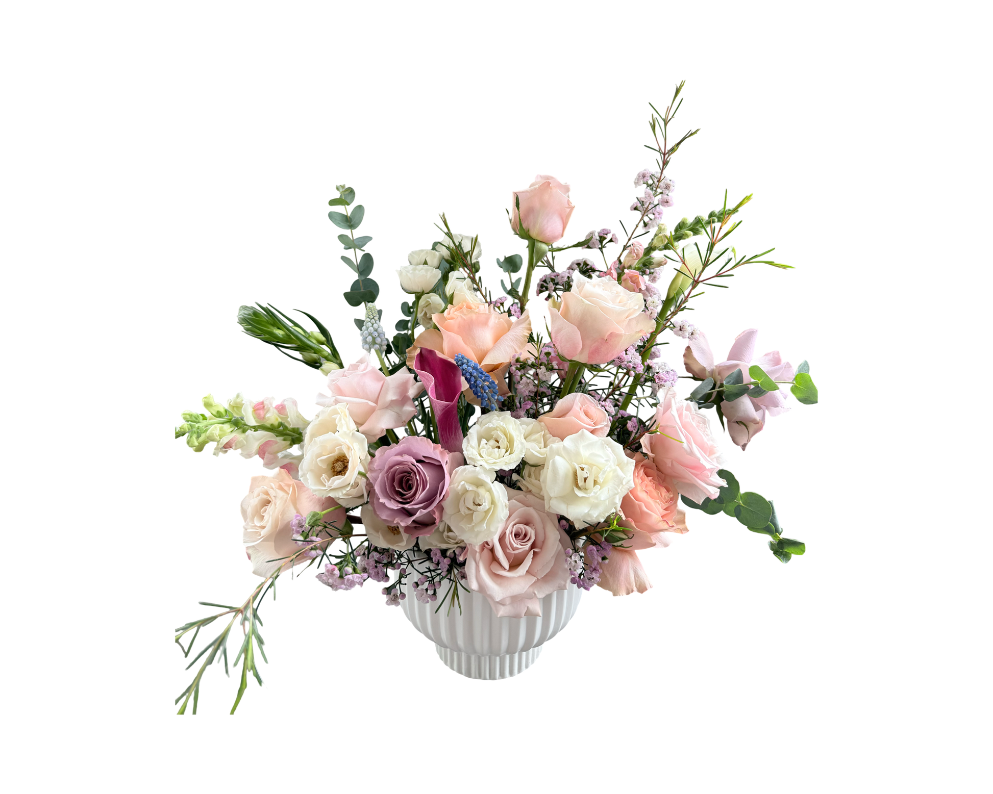 An ethereal garden-inspired floral arrangement designed in a fluted white ceramic vase, featuring soft blush roses, ivory spray roses, lavender roses, pale peach blooms, white ranunculus, pink waxflower, blue muscari, burgundy calla lilies, and touches of eucalyptus. With its natural flow and whimsical textures, this bouquet evokes a fresh spring meadow—perfect for refined gifting and serene occasions.