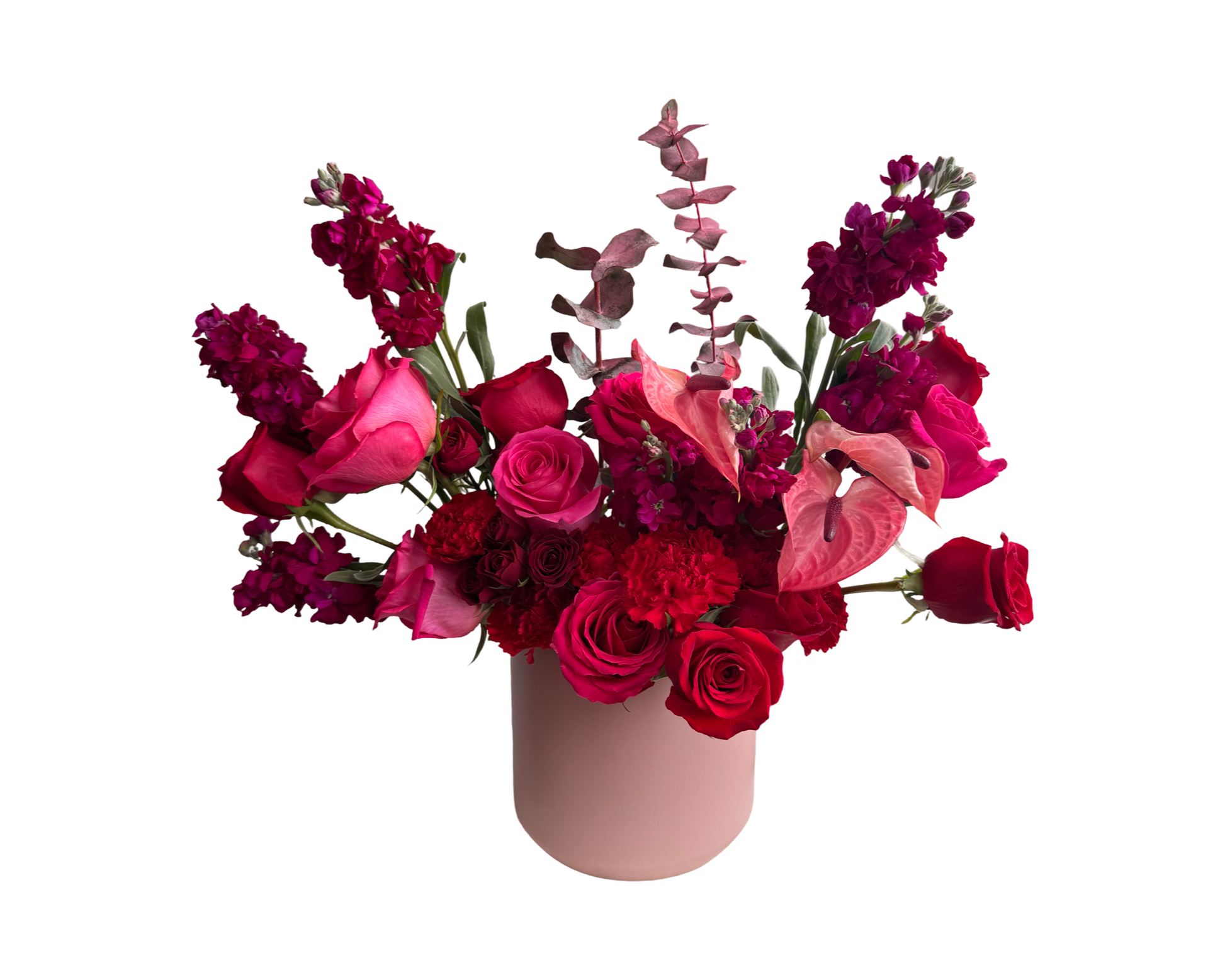 A striking floral arrangement in a matte blush pink vase, featuring hot pink roses, deep red roses, burgundy spray roses, red carnations, pink anthuriums, burgundy stock, and burgundy eucalyptus. The dramatic palette of fuchsia, crimson, and burgundy tones exudes passion and sophistication—ideal for Valentine’s Day, romantic gestures, or luxurious celebrations.
