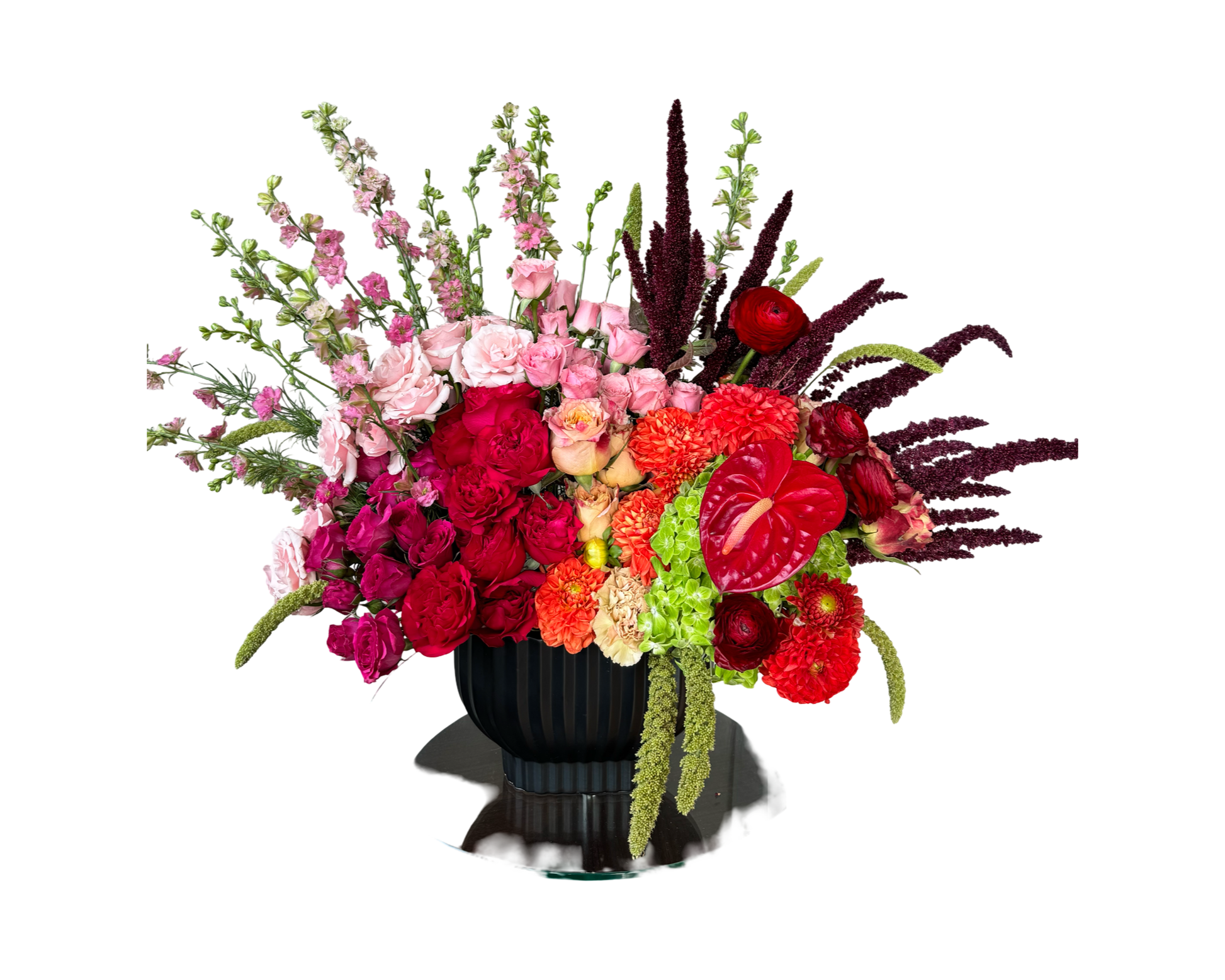 A dramatic and sculptural floral arrangement in a black pedestal vase, featuring a lavish blend of red ranunculus, pink spray roses, fuchsia roses, red anthuriums, orange carnations, lime green hydrangeas, blush roses, red amaranthus, and pink stock. With its asymmetrical design and vibrant palette, this arrangement makes a powerful statement perfect for luxury events, editorial styling, or unforgettable gifting.