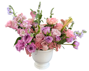 A romantic and elegant floral arrangement featuring blush pink roses, soft lavender roses, and graceful pink calla lilies. Delicate lavender stock flowers, lisianthus, and clusters of pink yarrow add texture and depth to the design. Arranged in a classic white vase, this pastel-toned composition is perfect for anniversaries, birthdays, proposals, Mother’s Day, or any occasion celebrating love and elegance.