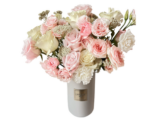 A soft and romantic floral arrangement presented in a tall white ceramic vase with a gold L’amour label. The bouquet features blush pink roses, ivory roses, white spray roses, baby’s breath, and white carnations. This airy and pastel-toned composition brings timeless elegance and tenderness, perfect for heartfelt gifting or meaningful celebrations.