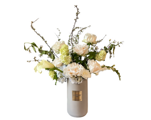 An elegant tall floral arrangement designed in a sleek beige L’Amour vase. It features soft ivory roses, lush hydrangeas, creamy calla lilies, and delicate green accent blooms. Wispy branches add graceful height and a natural, airy feel to the design. This arrangement blends classic whites and greens with subtle texture, making it a sophisticated choice for weddings, sympathy expressions, anniversaries, or refined home décor.