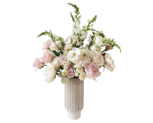 A tall and refined floral arrangement in a white fluted ceramic vase, featuring blush roses, white roses, ivory spray roses, lisianthus, white snapdragons, white peonies, and soft greenery. With a delicate pastel palette and timeless vertical silhouette, this bouquet is perfect for weddings, sympathy, baptisms, or romantic occasions.