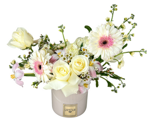 This delicate floral arrangement is set in a blush-toned, ribbed ceramic vase branded with "L’AMOUR". It features soft white and ivory roses, pale pink alstroemeria, white gerbera daisies with soft pink centers, small daisy-like chamomile blooms, and sprays of wax flowers. The color palette is fresh, clean, and softly romantic, ideal for spring or light-hearted occasions. Eucalyptus leaves and soft greenery offer texture and freshness, complementing the dainty, cheerful appearance.