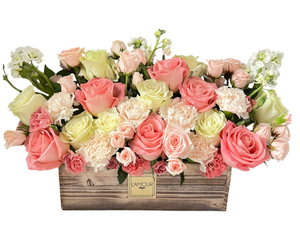 A soft and romantic floral arrangement featuring blush pink roses, creamy white roses, delicate spray roses, and a mix of pink and white carnations. Accented with clusters of stock flowers, the blooms are artfully arranged in a rustic wooden planter, giving the design a charming and elegant appeal. Perfect for anniversaries, birthdays, Mother’s Day, or as a thoughtful gift to brighten someone’s day with beauty and grace.