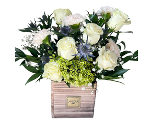 An elegant and calming floral arrangement featuring classic white roses, soft white carnations, and fresh green hydrangeas. Accents of striking blue thistle and delicate baby’s breath add texture and contrast, while lush greenery completes the look. Displayed in a rustic wooden planter with the L'amour Floral Designs label, this arrangement is perfect for anniversaries, birthdays, sympathy occasions, or as a refined centerpiece for any special event.