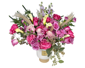 A bold and stylish floral arrangement featuring hot pink roses, soft pink roses, and lavender roses, paired with delicate spray roses and exotic calla lilies. Textured accents include pink carnations, airy pampas grass, and fresh eucalyptus, adding depth and visual interest. Presented in a chic white vase with a gold base, this modern arrangement is perfect for anniversaries, birthdays, or any occasion where a vibrant, elegant statement is desired.