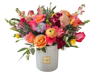 A colorful garden-style floral arrangement in a white ceramic vase from L’amour Designs, featuring a vibrant mix of orange roses, pink roses, yellow ranunculus, lavender scabiosa, yellow tulips, peach calla lilies, and blush stock. The varied textures and cheerful tones make this bouquet ideal for birthdays, celebrations, and brightening any occasion.