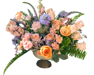 A soft and romantic floral arrangement featuring peach and blush roses, accented with vibrant orange roses and ranunculus. Light peach spray roses and carnations add ruffled texture, while lavender scabiosa blooms bring contrast and charm. Accents of veronica, greenery, and ferns create height and a natural flow. Arranged in a vintage-style pedestal vase, this design exudes elegance and grace, perfect for anniversaries, birthdays, or Mother’s Day celebrations.