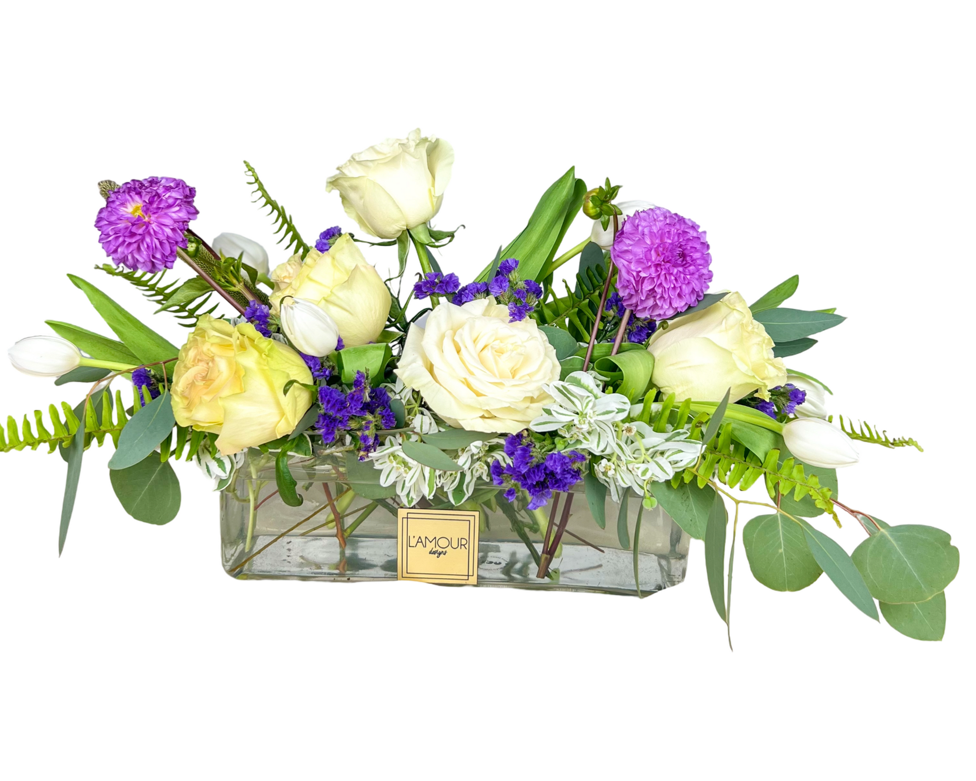 This modern floral arrangement is crafted in a sleek rectangular glass vase, combining creamy white roses, ivory tulips, yellow roses, and bold pops of purple chrysanthemums and statice. Greenery and ferns are artfully woven through the design to create a fresh, elegant, and cheerful centerpiece. Ideal for celebrations, office decor, or a sophisticated gift.
