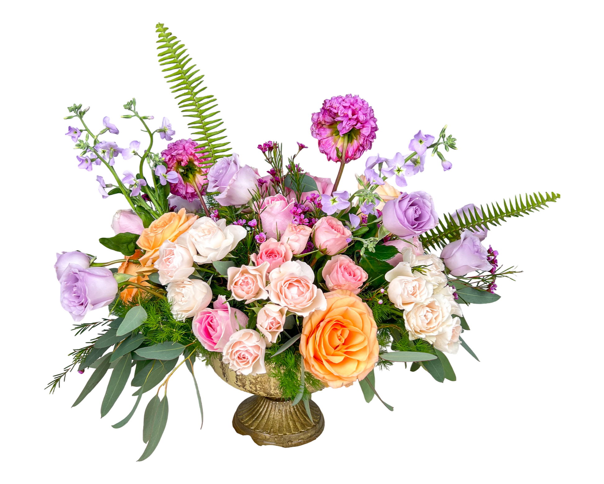 A garden-inspired floral arrangement in a vintage gold pedestal vase, bursting with lavender roses, peach and blush spray roses, creamy white roses, pink waxflower, deep magenta carnations, purple stock, and lush greenery including eucalyptus and ferns. This airy and artistic bouquet exudes a graceful charm and romantic feel, ideal for spring celebrations and elevated gifting.