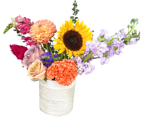 A cheerful and vibrant floral arrangement featuring a sunny yellow sunflower, warm peach and lavender roses, soft lilac stock flowers, peach dahlias, and textured orange and magenta celosia. Accents of deep purple statice and fresh eucalyptus add depth and contrast. Arranged in a modern white and beige textured vase, this lively mix of blooms exudes happiness and warmth, making it an ideal gift for birthdays, get well wishes, or to brighten any home or office space.