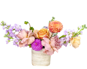 A charming pastel floral arrangement featuring soft peach roses, blush lavender roses, and vibrant dahlias in shades of coral orange and vivid magenta. Delicate lavender stock flowers extend gracefully, adding texture and height to the composition. Arranged in a textured white vase, this elegant yet cheerful design is perfect for birthdays, anniversaries, housewarmings, or as a thoughtful pick-me-up to brighten someone’s day.