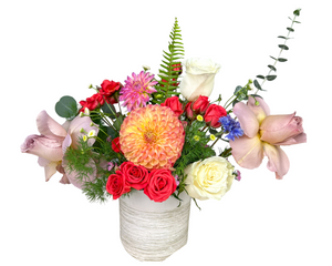 A colorful and charming floral arrangement featuring a large peach dahlia as the focal bloom, paired with soft lavender roses, creamy white roses, and clusters of vibrant red spray roses. Accents of purple dahlias, blue cornflowers, and delicate wax flowers add pops of contrast, while ferns and eucalyptus lend a fresh, natural balance. Arranged in a modern white vase, this lively bouquet is perfect for birthdays, anniversaries, or Mother’s Day celebrations.