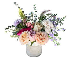 A delicate garden-style arrangement in a textured taupe ceramic vase, featuring a soft mix of peach and blush roses, lavender spray roses, pink spray roses, lavender aster, bunny tail grass, eucalyptus, and light greenery. The fresh, romantic color palette makes it perfect for spring gifting, baby showers, or everyday elegance.