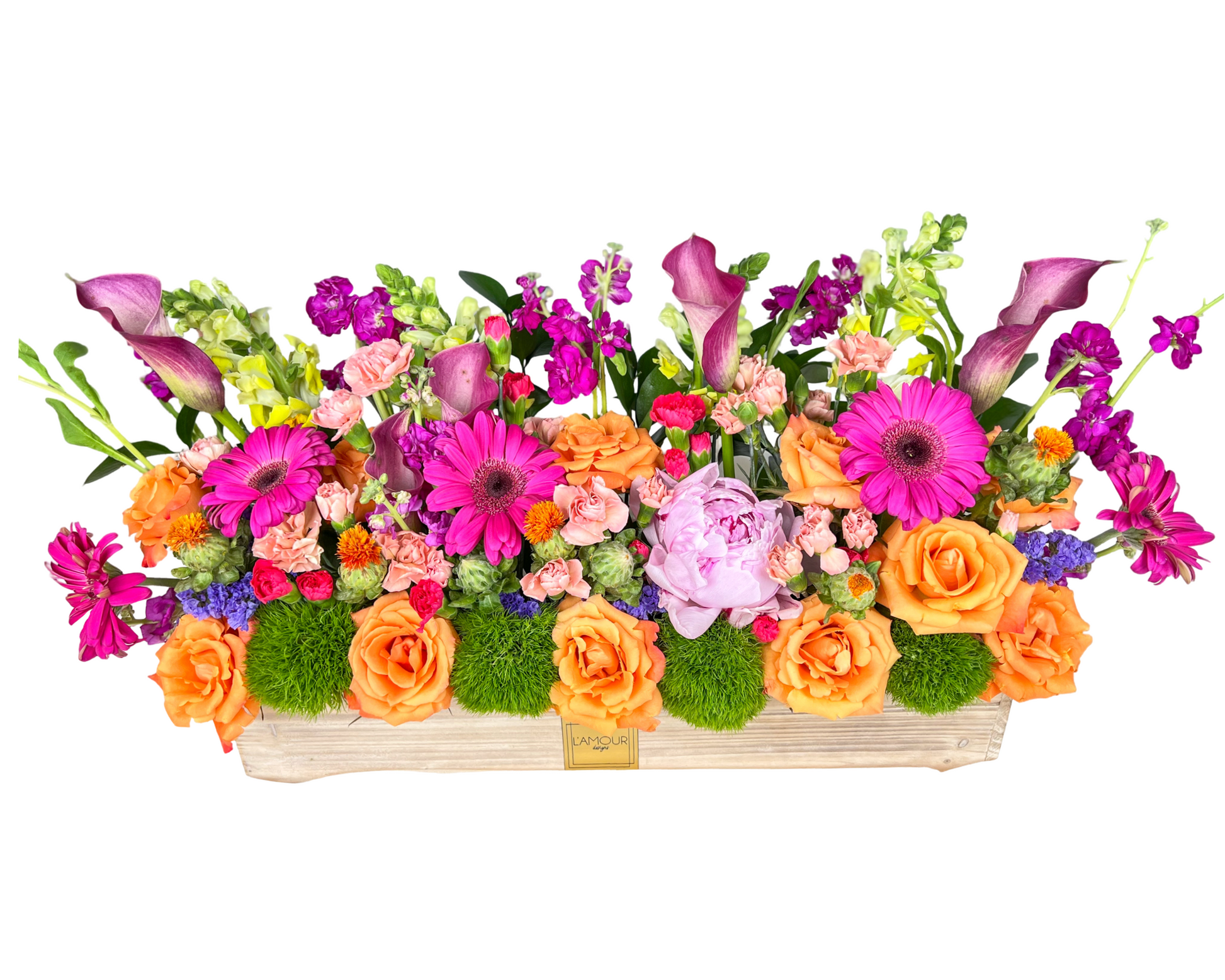 A lively and colorful rectangular floral centerpiece arranged in a light wooden L’amour Designs planter. This vibrant design includes orange roses, hot pink gerbera daisies, lavender calla lilies, pale pink carnations, purple stock, yellow snapdragons, pink peonies, lime green moss balls, purple statice, and assorted wildflower accents. A cheerful and celebratory piece perfect for festive gatherings or joyful gifting.
