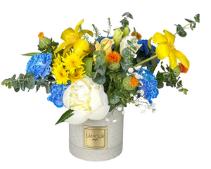 A bright and refreshing floral arrangement featuring cheerful yellow irises, sunny yellow chrysanthemums, and soft white peonies. Accents of vibrant blue carnations and unique orange safflowers add contrast and texture. Baby’s breath and eucalyptus greenery provide a light, airy touch, all beautifully arranged in a modern white vase with the L'amour Floral Designs label. This lively design is perfect for birthdays, anniversaries, housewarmings, or as a joyful gift to brighten any space.