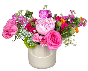 A vibrant and colorful floral arrangement featuring hot pink roses, lush pink peonies, and soft pink carnations. Accents of spray roses, purple stock flowers, bright green chrysanthemums, delicate baby’s breath, and unique orange safflower add texture and contrast. Presented in a modern white ribbed vase, this arrangement is perfect for birthdays, anniversaries, Mother’s Day, or as a joyful gift to brighten someone’s space.