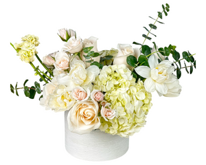 A delicate and elegant floral arrangement in a white ceramic vase, featuring ivory roses, blush spray roses, white hydrangeas, creamy lisianthus, white tulips, yellow stock, and eucalyptus. This pastel and ivory-toned piece is perfect for weddings, sympathy, baby showers, or everyday luxury gifting.