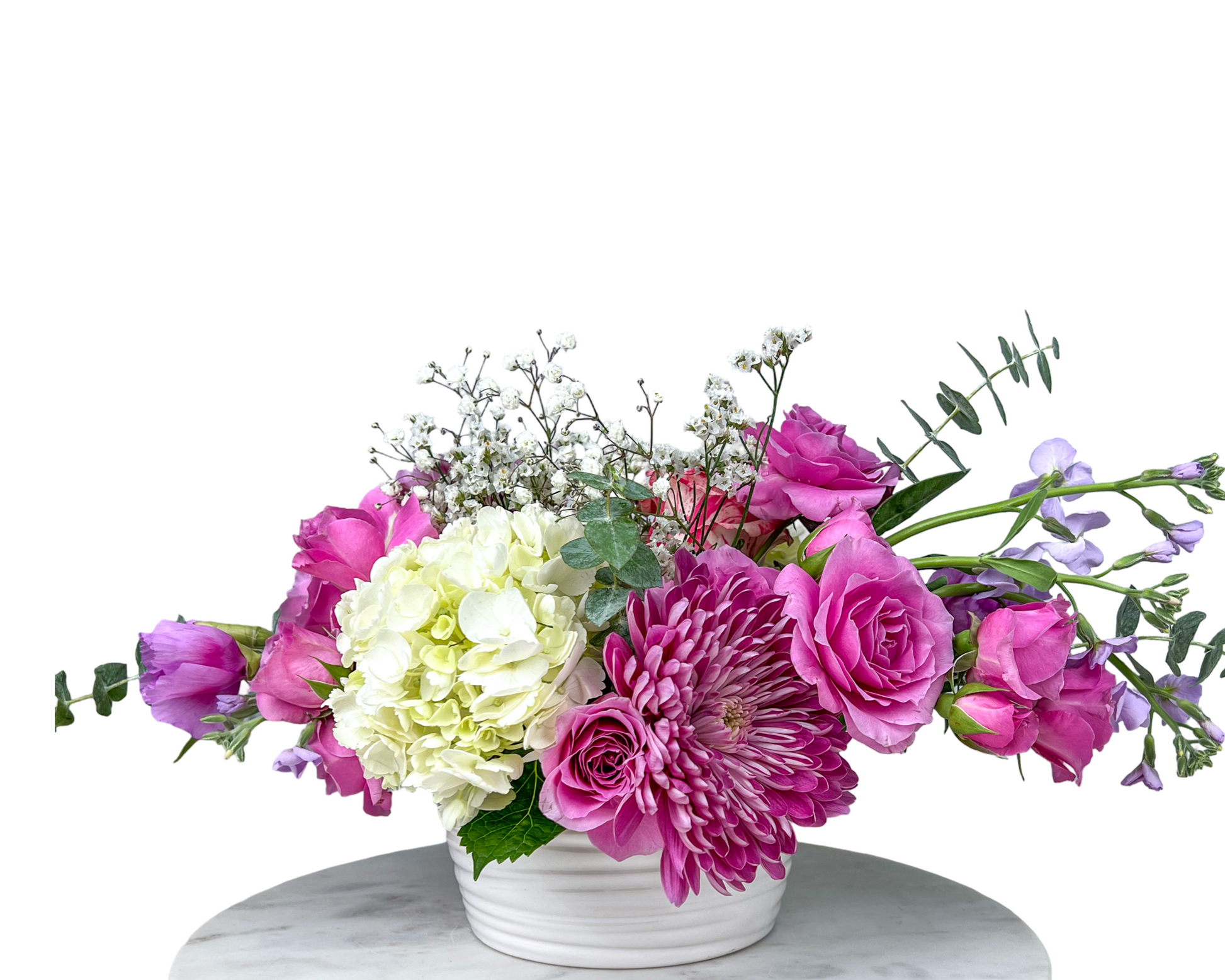 This floral arrangement features a bright and cheerful palette of pink and lavender tones. Prominent blooms include lavender roses, soft purple lisianthus, white hydrangeas, bold pink chrysanthemums, and baby's breath. Eucalyptus and greenery are used as fillers, adding texture and natural balance. The flowers are arranged in a white ceramic vase placed on a marble surface, giving the entire design a modern yet elegant look.