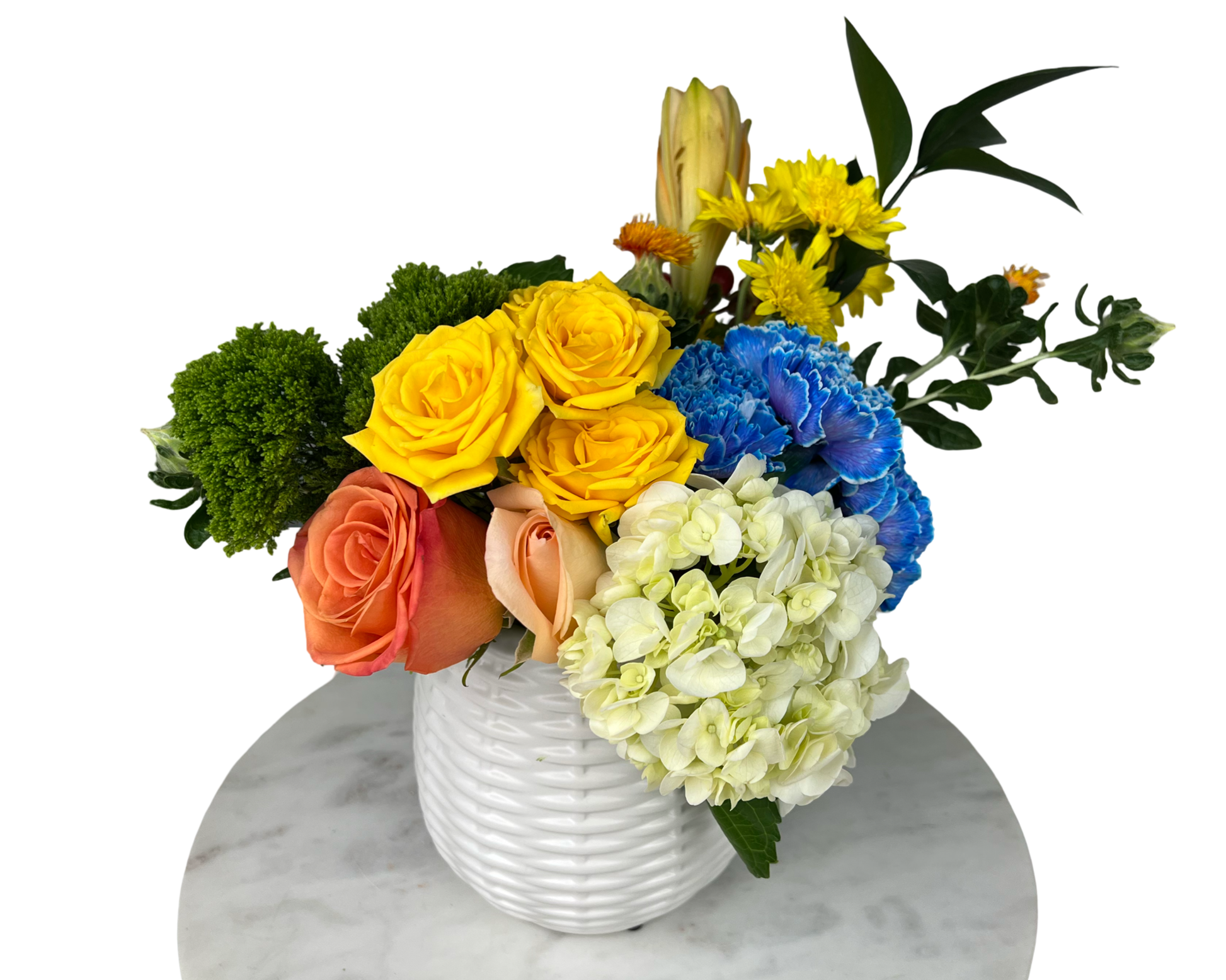 A compact and colorful floral arrangement in a textured white ceramic vase, featuring yellow roses, orange and peach roses, vibrant blue carnations, white hydrangeas, yellow mums, green moss accents, and tropical foliage. This playful and high-contrast bouquet is perfect for brightening up any occasion, offering a unique and celebratory look.