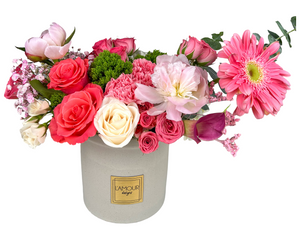 This floral arrangement features a vibrant mix of pink and fuchsia blooms in a light gray ceramic vase labeled "L’Amour Designs." It includes vivid hot pink gerbera daisies, soft pink peonies, classic pink and cream roses, calla lilies, carnations, tulips, and baby’s breath, creating a lush and romantic display. The contrasting textures of the various blooms and soft greenery add visual interest and dimension, making it a cheerful and charming bouquet perfect for celebrations or heartfelt gestures.